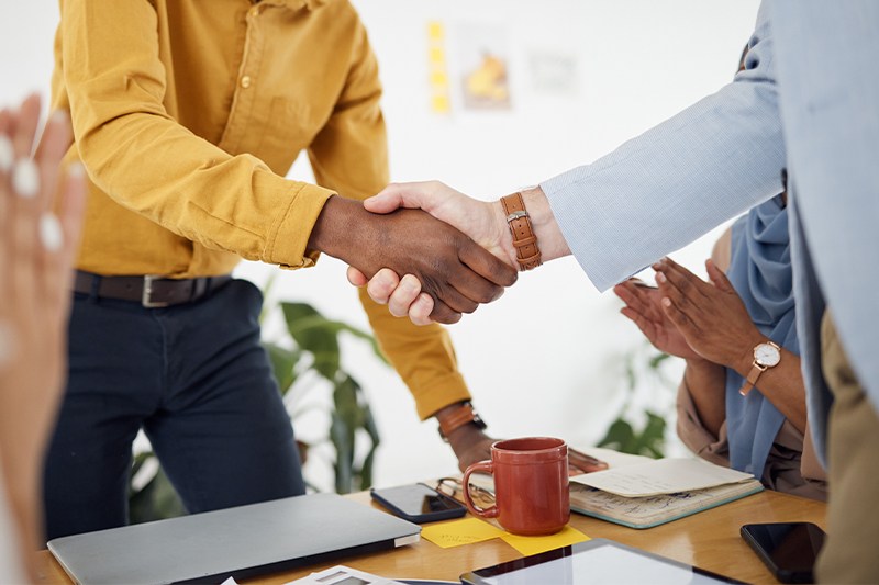 Two professionals shaking hands while colleagues clap around them